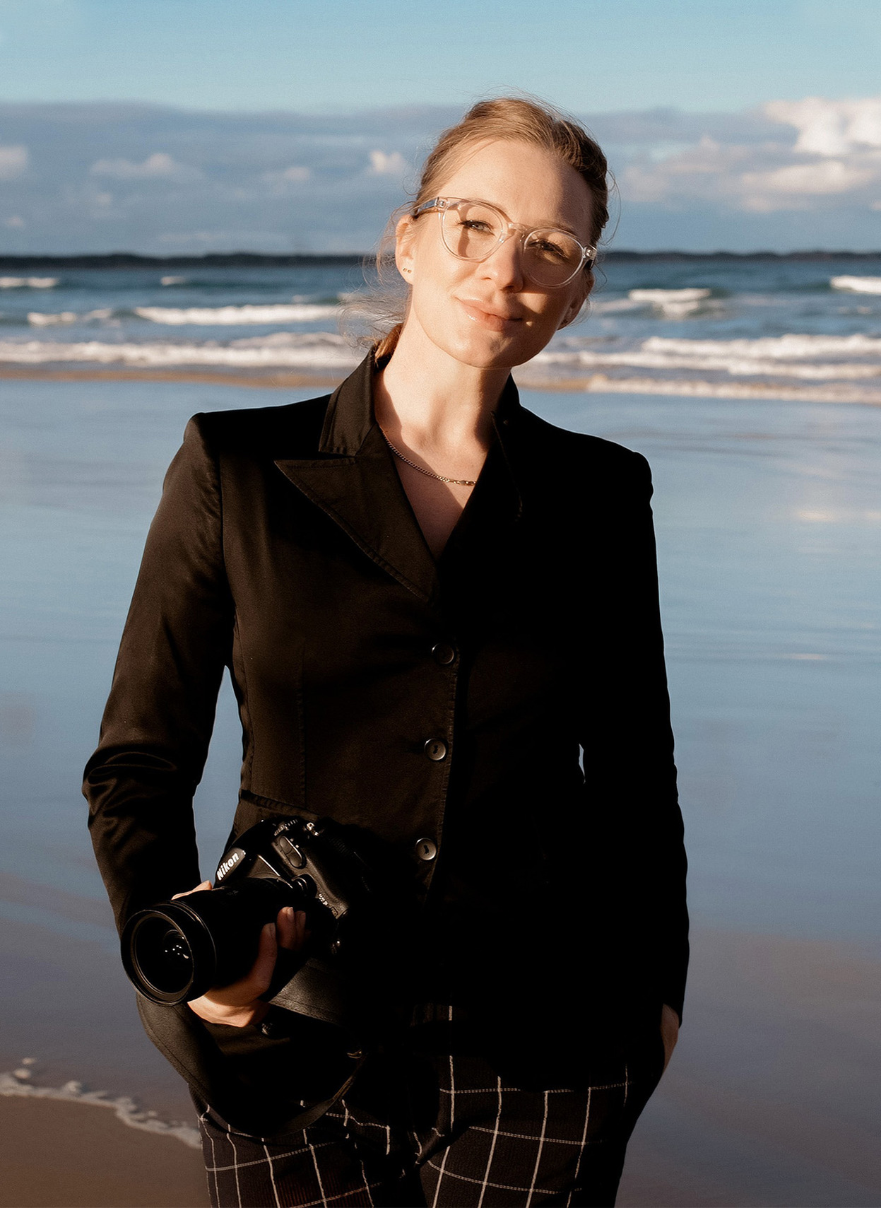 Melbourne wedding female photographer smiling while holding a camera, wearing all black. The background features a wet shoreline and the sea.