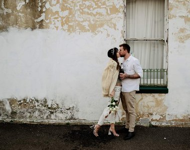 male and female kissing in front of old worn side of house with window
