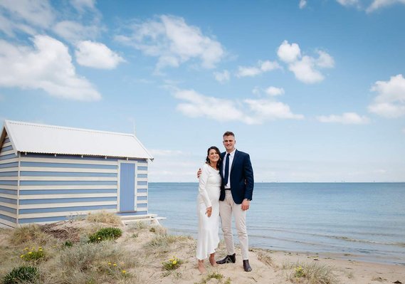 Man in suite holding lady by her waist. Lady wearing white. cabin in the background, posing with the beach and sky.
