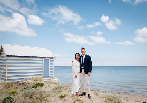 Man in suite holding lady by her waist. Lady wearing white. beach cabin in the background, posing with the beach and sky.
