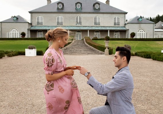 Man in suite and woman in pink dress holding, man is on one knee, double story building behind them.