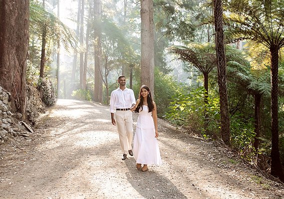 Male and female walking on a path in a forest with the sunlight beaming down behind them. Girl wearing a white dress. Man wearing white shirt and tan pants.