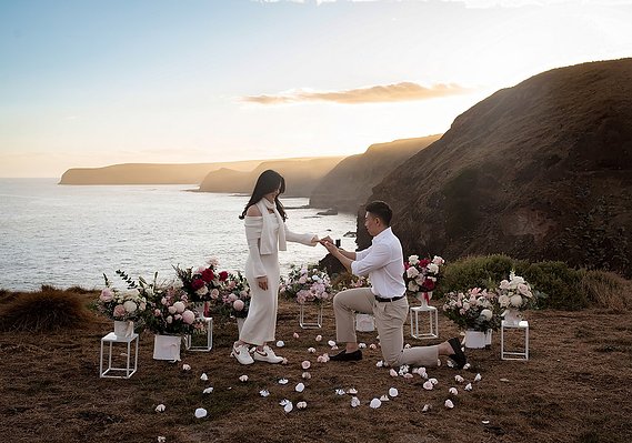 male proposing to female on cliff with views of ocean. flower bouquets set up around the couple.