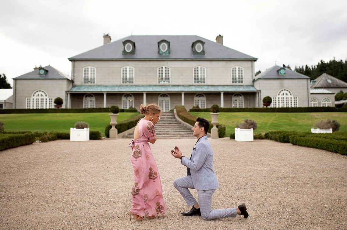 Man on one knee, woman in pink dress with hands on face. house in back ground