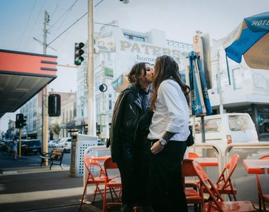 Male wearing a white shirt, kissing his female partner, who is wearing a shiny, dark blue dress. Red tables and chairs are in the background, with a city scene behind them.