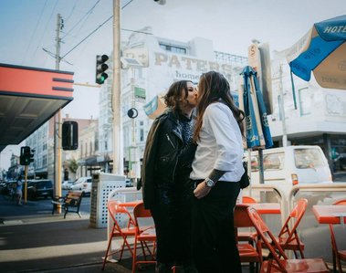 Male wearing a white shirt, kissing his female partner, who is wearing a shiny, dark blue dress. Red tables and chairs are in the background, with a city scene behind them.