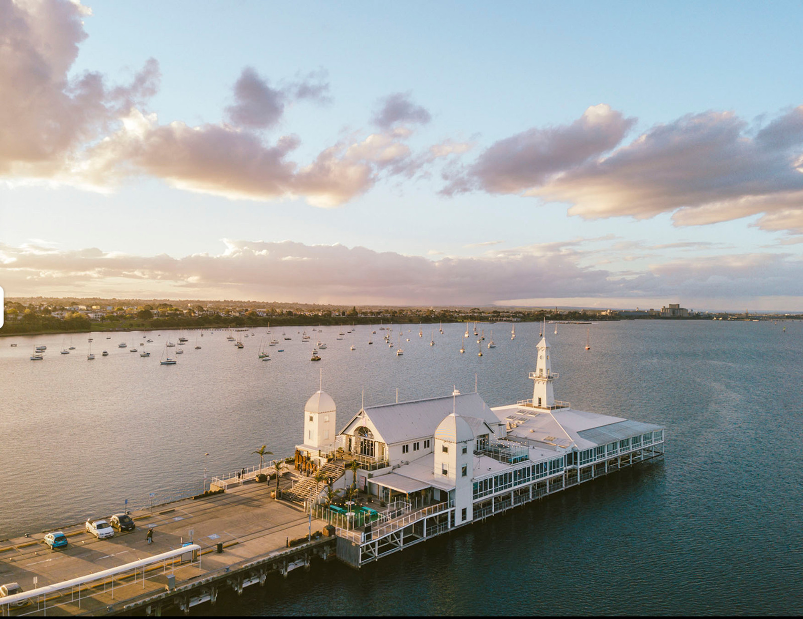 A large white building, with chairs and tables on the outside, at the end of a pier. The background features a sunsetting shoreline, clouds and the sea.