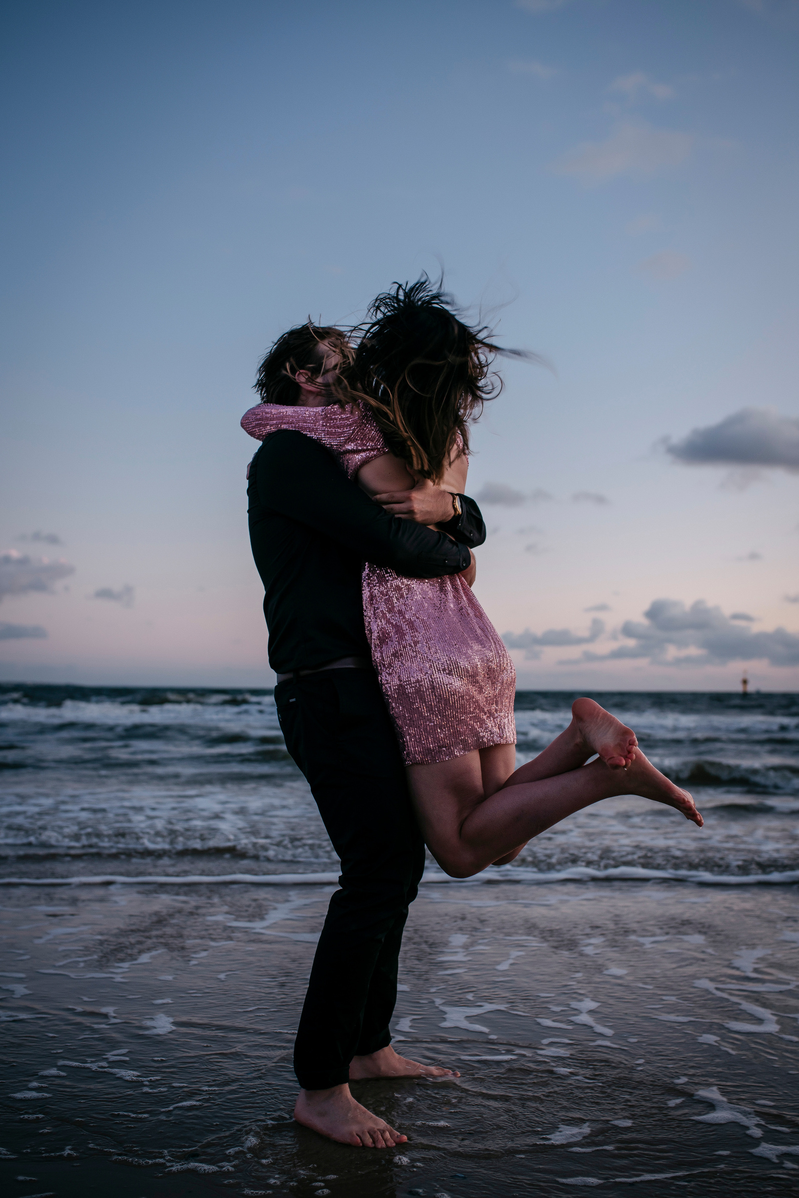 A woman jumping into the arms of her partner on the shore, with the sea in the background.