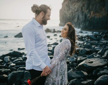 Male and female couple, both wearing white, stood on a rocky shoreline while smiling at each other. The sea is directly behind them.