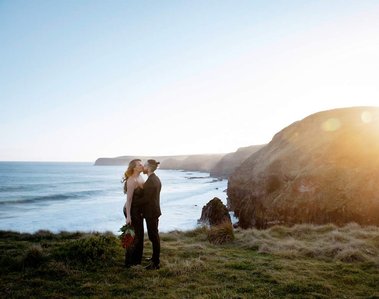 Male and female couple, both wearing black, kissing with a view of the cliffs and sea in the background.