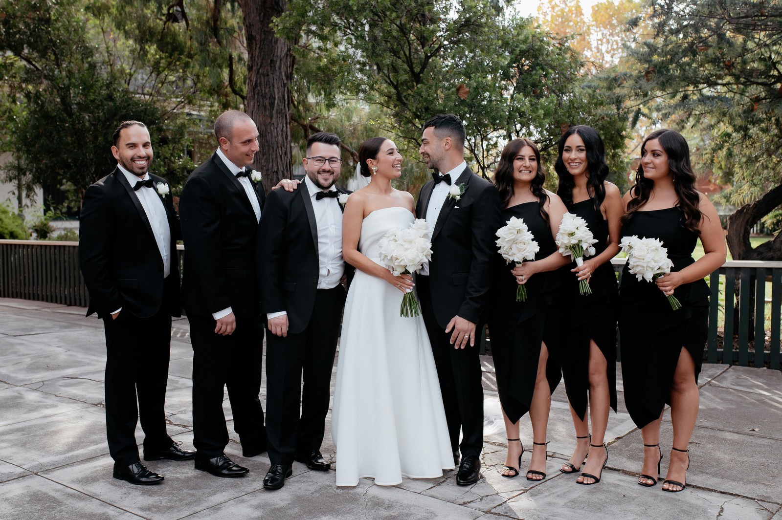  3 groomsmen and 3 bridesmaids, all wearing black, stood alongside a male and female partner. The male is in a black suit, while the woman is in an all-white wedding dress. The bridesmaids and bride are holding white flower bouquets.