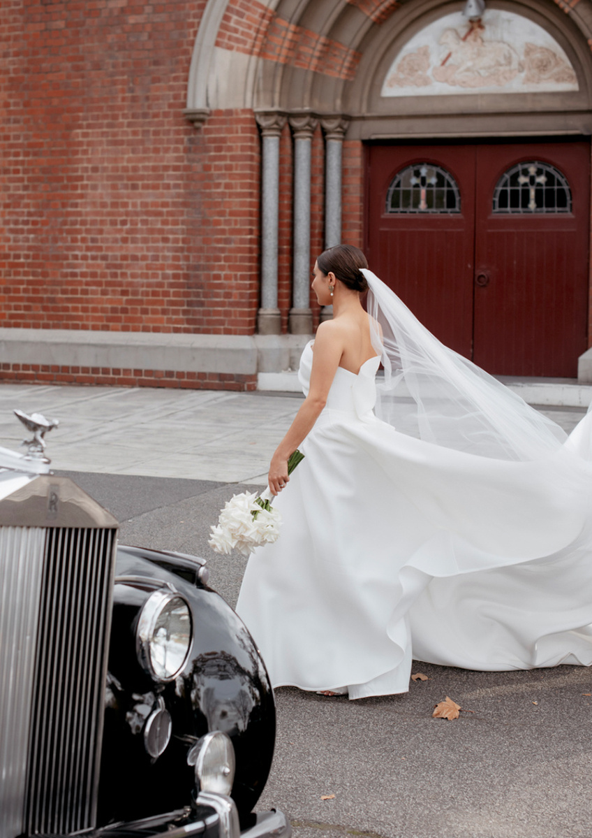 Bride walking past a church with vintage car in foreground