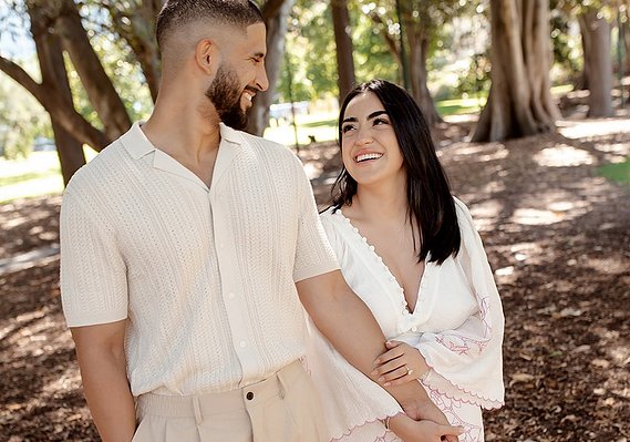 male and female walking hand in hand looking at each other. walking in garden area with large trees in background. 