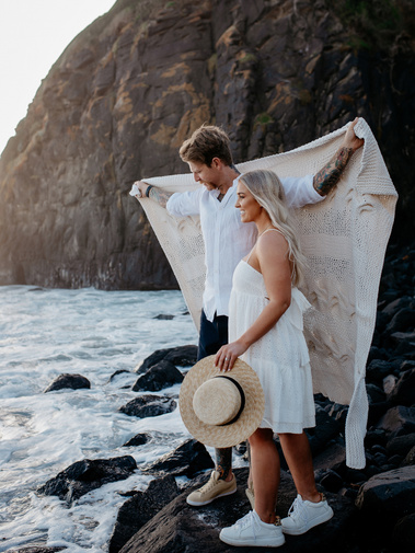 Male holding a beige blanket behind the back of him and his female partner. The pair are stood on a rocky coastline, next to the sea.