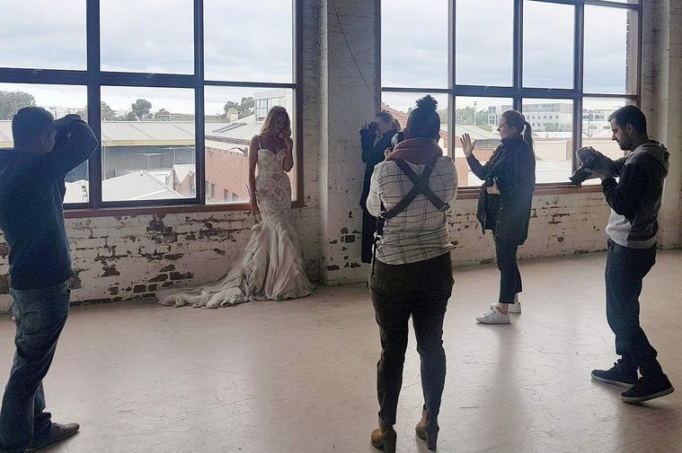 5 photographers photographing bride in dress. Bride in front of large warehouse windows. 