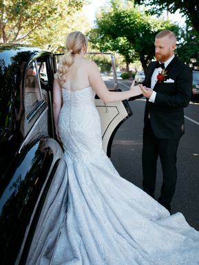 Woman wearing a white wedding dress, with her male partner holding her hand as she enters a luxury car.
