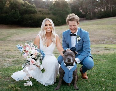 Woman wearing a white wedding dress, holding a bouquet of flowers, kneeling next to her partner who is wearing a blue and white suit with black bow tie. They are both holding onto their dog, who is wearing a similar suit to the male.