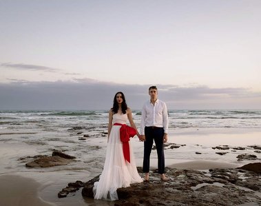 Lady in white dress with big red bow, man in white shirt behind her. beach and rocks 
