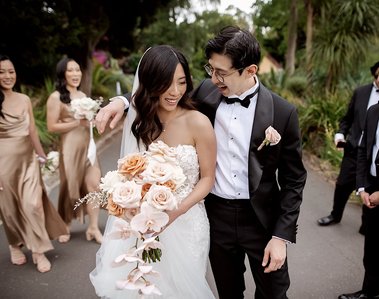 Groom with arm around brides shoulder walking looking each other in park setting with bridal party in the background. 