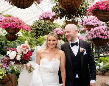 Flowers hung on ceiling and in background. man in black suite and woman in a white wedding dress are walking. woman has flowers in her hand. 