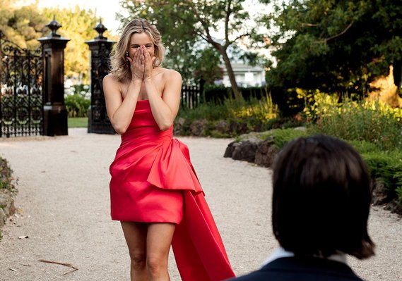 Lady in red dress hands on her face standing, man with long hair from behind