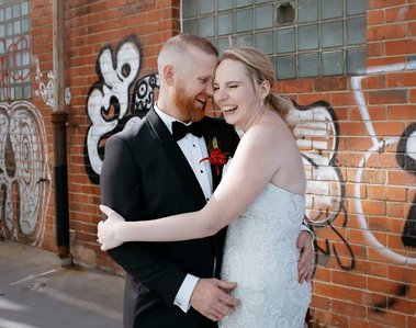 Bride and groom in industrial brick setting with graffiti embracing each other with eyes closed laughing. 