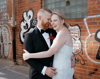 Man holding his female partner by the waist, side on to the camera. The background features a graffitied brick wall.