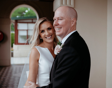  Man wearing a black and white holding his female partner, who is wearing a white wedding dress. The woman is looking at the camera and smiling, while the man looks into the distance, towards the left of the picture.
