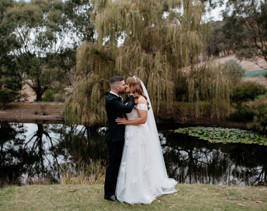 Woman in a white wedding dress, kissing her partner who is in a black suit. The couple are stood in front of a lake, with a tree hovering in the centre.