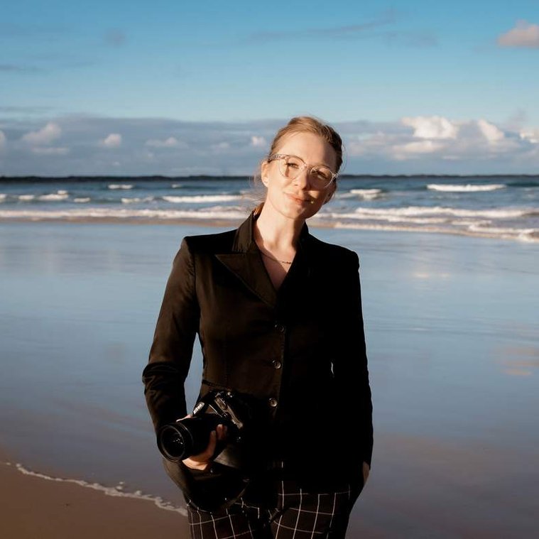 Women holding camera  in hand, looking at the camera, wearing black blazer with beach in background