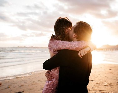 Man holding his female partner in the air while kissing her. The male is wearing a black shirt, while the woman is wearing a pink, sequin dress. They are stood on a beach with the sea behind them.