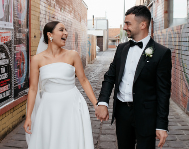 Female in a white wedding dress holding her male partners' hand. The male is wearing a black and white suit, with a Melbourne laneway as the background.