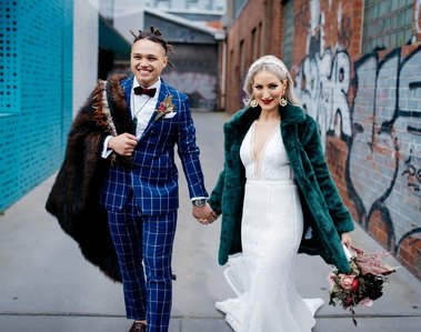 an wearing a blue checkered suit with white shirt and blue bow tie, holding his female partners hand while they both walk towards the camera. The woman is wearing a white wedding dress with a green furr coat and is holding a bouquet of flowers.