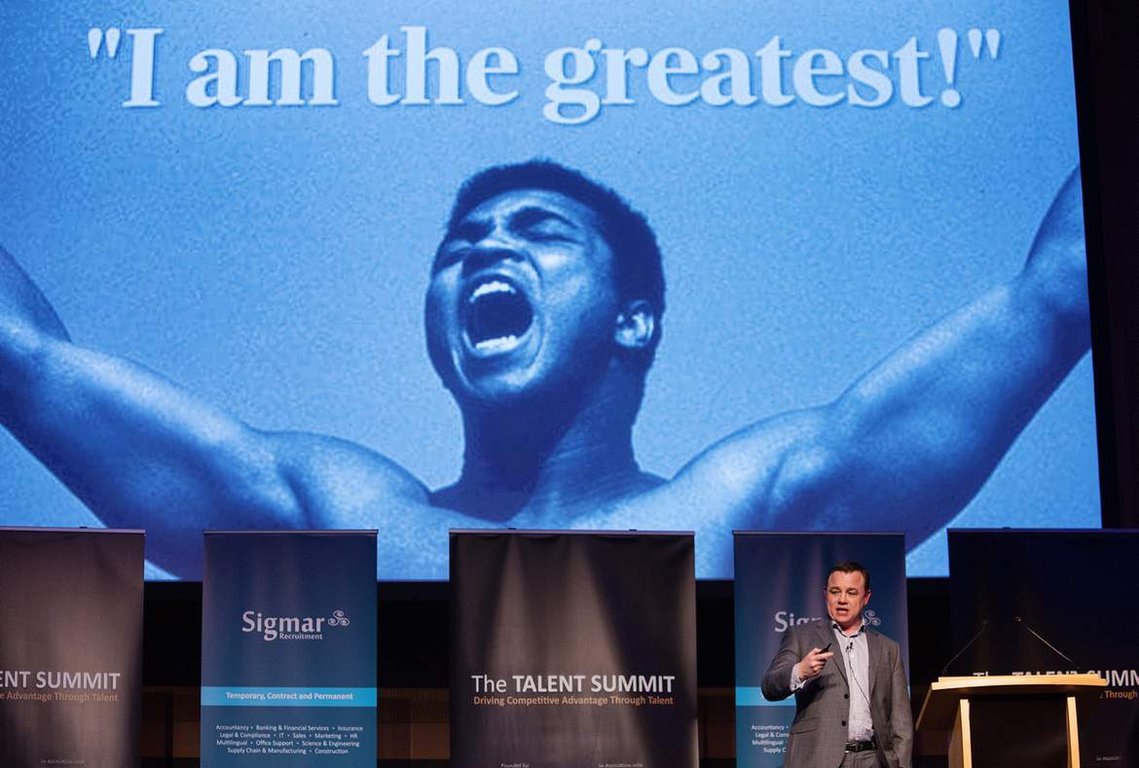 Corporate photo of male speaker presenting at an international conference in Dublin with muhammad ali 'I am the greatest' image in background. Live Event Photography