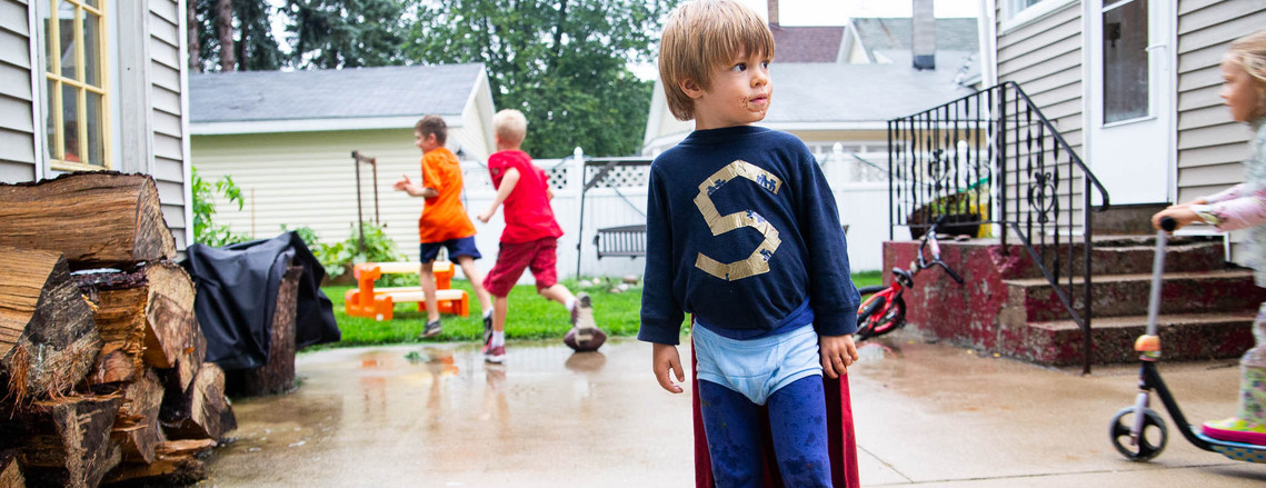 A young child playing dress up in what looks like a homemade superman costume with kids running around in the background.