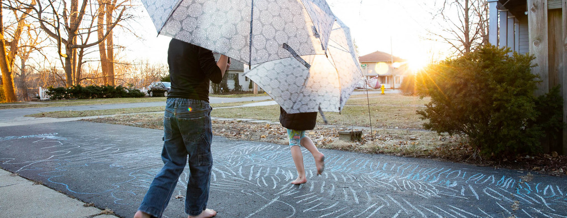 Two children with umbrellas dancing in the sun on a driveway covered in sidewalk chalk