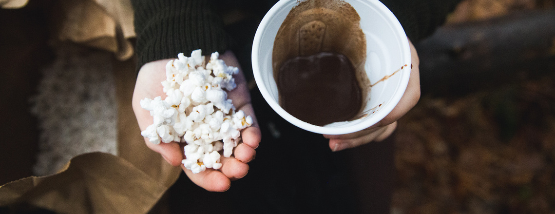 One hand is holding a handful of popcorn and the other is a cup of hot chocolate.