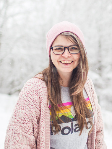 Portrait of a woman wearing a pink hat, glasses, a polaroid brand t-shirt and a pink sweater with a snow covered background.