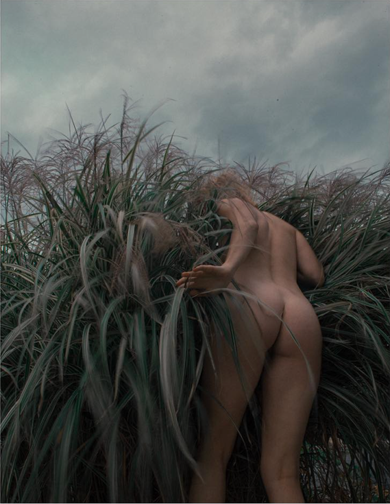 Nude woman lunges into tall grass, grey cloudy skies
