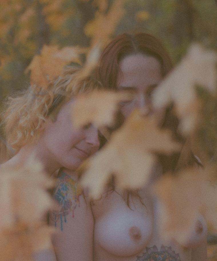 Vintage film photo of two nude women embracing behind yellow autumn maple leaves in the woods