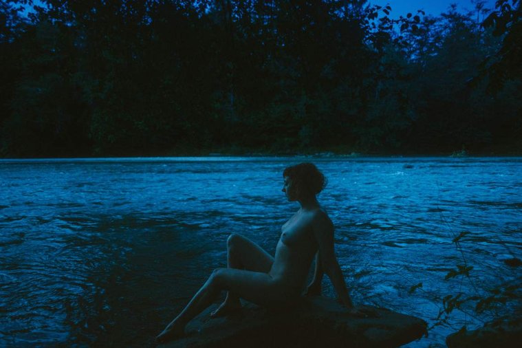Nude woman lounges on rock by river against blue sky at dusk in Olympic National Park, Washington