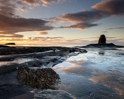 Sunset clouds reflected in the shallow water of Saltwick Bay, with Black Nab in the background. A photograph by Tim Pearson.