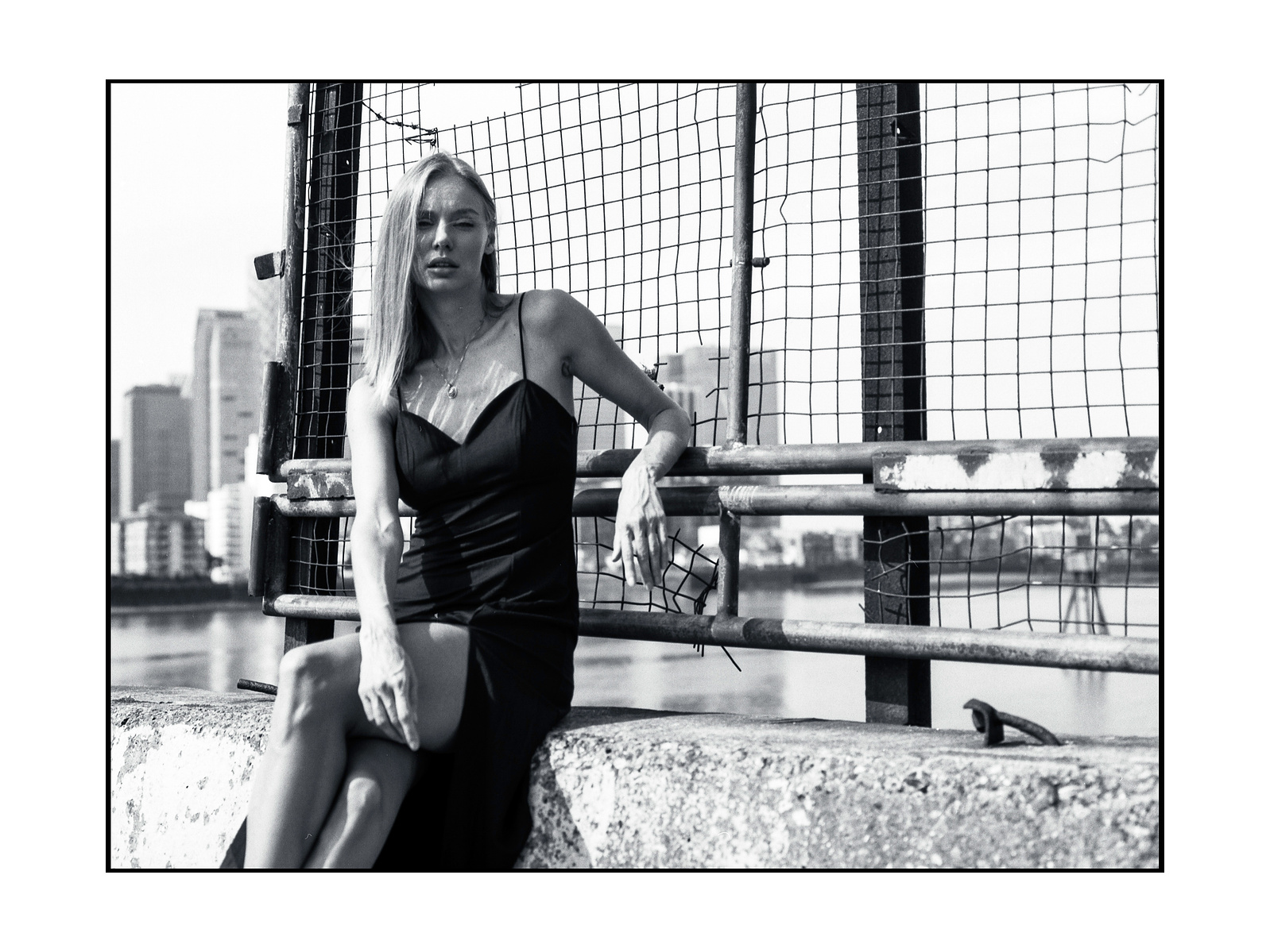 Woman seated by some railings and a fence. arm resting on the fence via elbow wearing black dress.