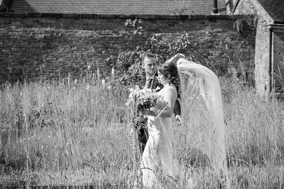 relaxed and natural wedding photography, black and white image of bride and groom walking through long grass. bride messing with veil that is blowing in wind