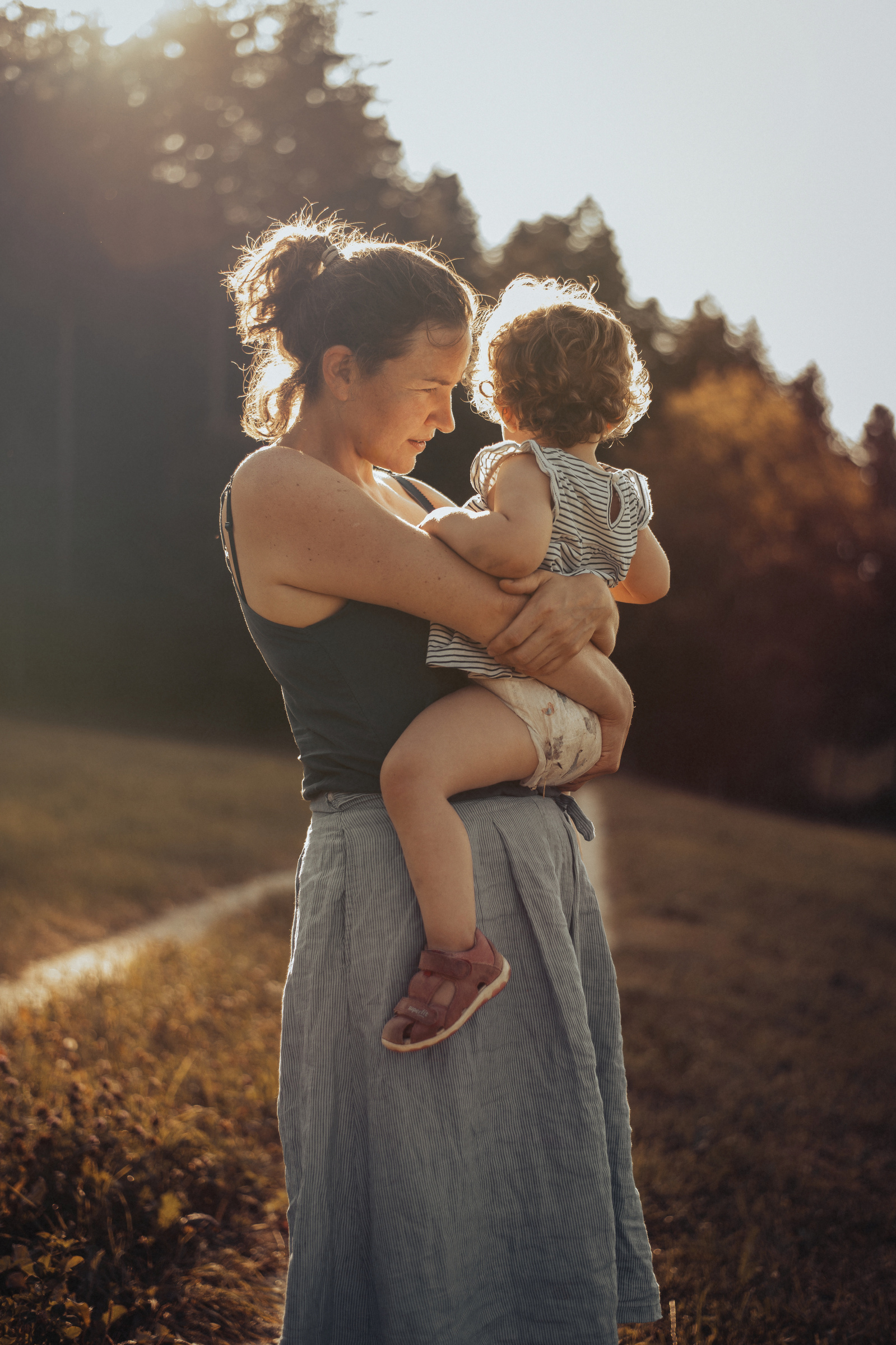 Junge Frau mit ihrem Kind auf dem Arm in der stimmungsvollen Abendsonne auf einer Wiese stehend.
