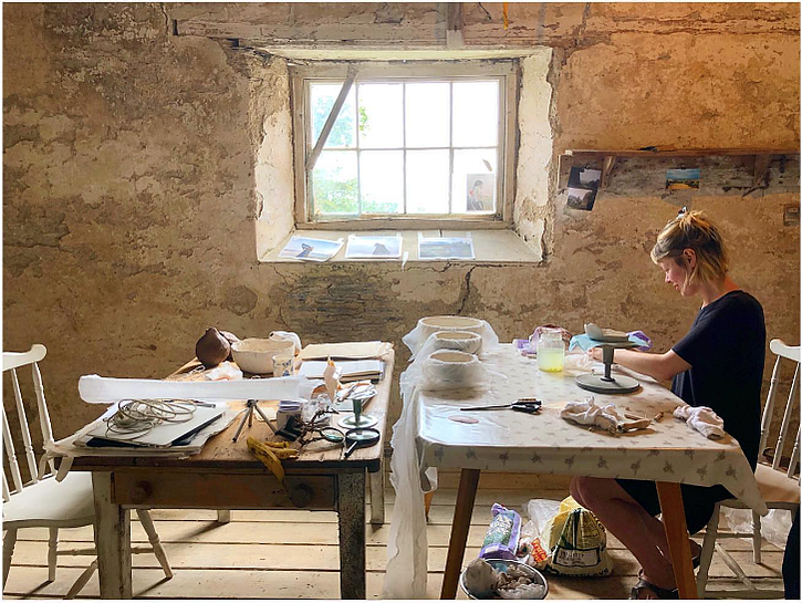 Alice is sitting at a desk in a ceramics studio with rustic plaster walls and a window. She is a white female wearing a black dress.