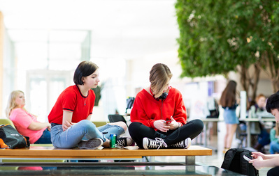 Two high schools wearing red are sitting cross legged are sitting on a bench at the Discovery building on the University of Wisconsin Madison campus. 