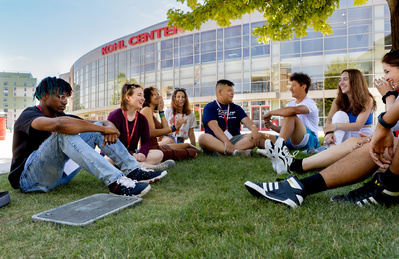A group of kids sit in the grass outside the Kohl Center at the University of Wisconsin. It's a nice summer day. They are stoked. Maybe. 