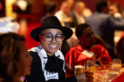 A stylish women wearing a hat is sitting a table and smiling while talking to someone. She's at the Monona Terrace. 