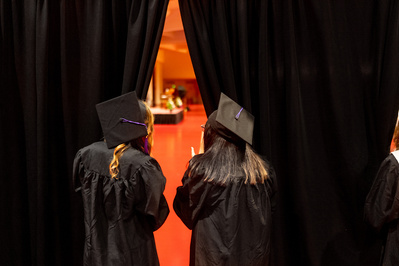 Two graduates are looking through a curtain at the ceremony happening behind the curtain. 